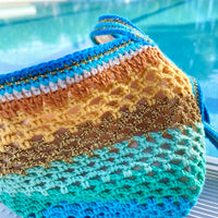 Paris designed Handmade crochet bag seen from the front, gradient of electric blue, terracotta, yellow, and turquoise, with gold highlights, in front of a swimming pool. Detailed view of the colors and stitch.
