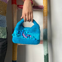 femme debout portant un haut en crochet Anabaum et à la main le sac en crochet Anabaum corail Turquoise dans les tons bleu canard et turquoise, texturé comme du corail