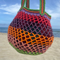 Lil Sis Funky crochet mesh bag held up against a blue sky on the beach, vibrant green, orange and pink stripes, designed in Paris by Anabaum