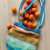 anabaum OG Sirena crochet market bag filled with apricots, laid flat on a wooden surface with blue handles