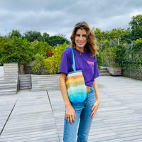 Woman standing on a wooden deck wearing a purple top and jeans, holding an anabaum sirena og bag, rainbow gradient crochet shoulder bag
