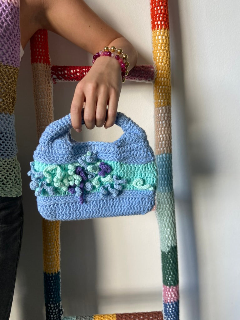 femme debout portant un haut en crochet Anabaum et à la main le sac  en crochet Anabaum Bleu Ciel dans les tons Bleu ciel céladon , texturé comme du corail 
