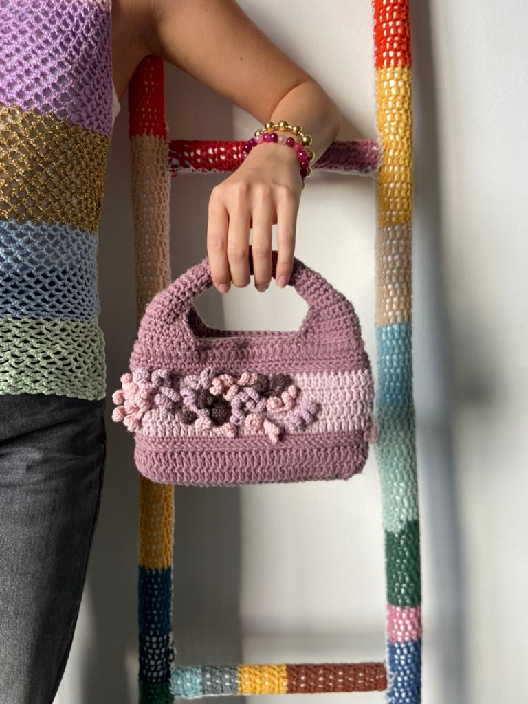femme debout portant un haut en crochet Anabaum et à la main le sac  en crochet Anabaum Corail Parme dans les tons parme et rose pale , texturé comme du corail 