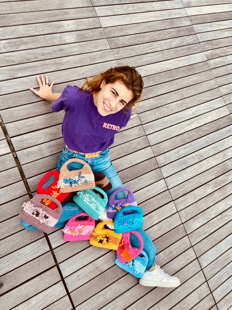 femme souriante assise par terre regardant l'objectif, vue de haut avec sur ses jambes  une douzaine de sacs au crochet Anabaum Corail tous de différentes couleurs vives