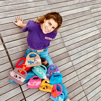 femme souriante assise par terre regardant l'objectif, vue de haut avec sur ses jambes  une douzaine de sacs au crochet Anabaum Corail tous de différentes couleurs vives