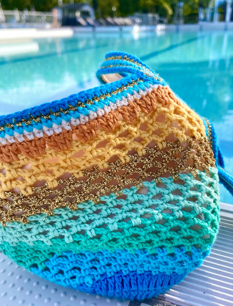 Paris designed Handmade crochet bag seen from the front, gradient of electric blue, terracotta, yellow, and turquoise, with gold highlights, in front of a swimming pool. Detailed view of the colors and stitch.