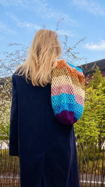 Femme de dos marchant sur un muret au bord d’un bassin, portant un long manteau bleu et un jean, avec un grand sac crochet multicolore Anabaum , le Maxi Cosmic, sur l’épaule, sous un ciel bleu printanier.