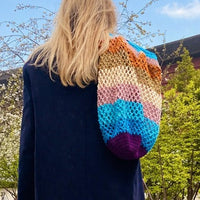 Femme de dos marchant sur un muret au bord d’un bassin, portant un long manteau bleu et un jean, avec un grand sac crochet multicolore Anabaum , le Maxi Cosmic, sur l’épaule, sous un ciel bleu printanier.
