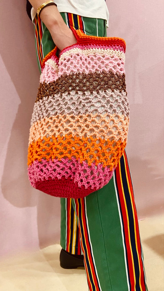 Sac cabas en crochet multicolore Anabaum , Maxi Vibe, porté à la main, avec rayures dégradées rose, orange, beige et marron, associé à un pantalon vert à rayures colorées et tshirt blanc.