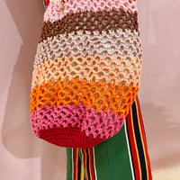 Sac cabas en crochet multicolore Anabaum , Maxi Vibe, porté à la main, avec rayures dégradées rose, orange, beige et marron, associé à un pantalon vert à rayures colorées et tshirt blanc.
