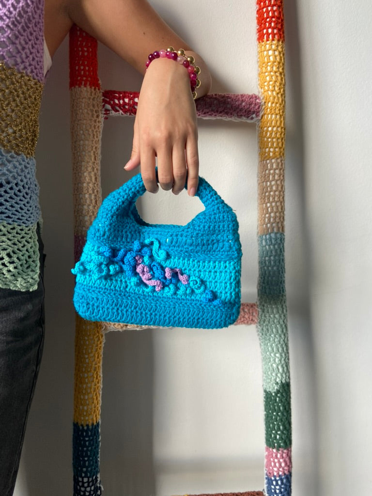 femme debout portant un haut en crochet Anabaum et à la main le sac  en crochet Anabaum corail Turquoise dans les tons bleu canard et turquoise, texturé comme du corail 