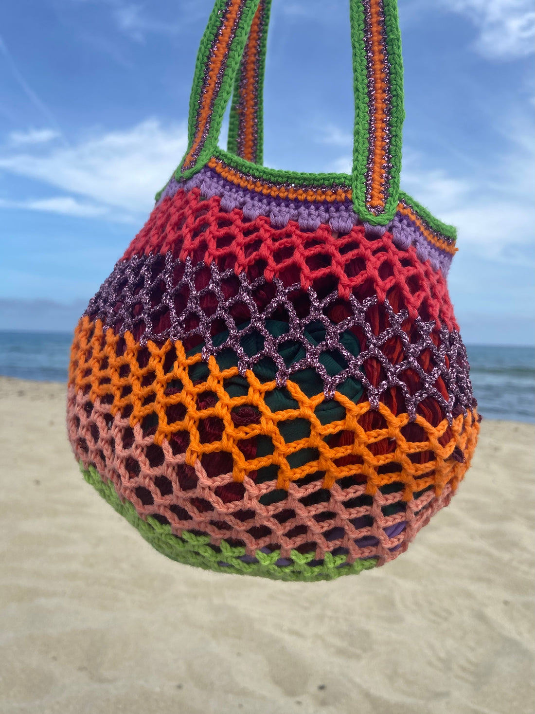 Lil Sis Funky crochet mesh bag held up against a blue sky on the beach, vibrant green, orange and pink stripes, designed in Paris by Anabaum