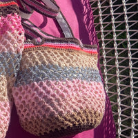 Close-up of the Lil Sis Sandstorm handmade crochet bag in pink, mauve and grey stripes, resting on a wicker chair with a fuchsia background