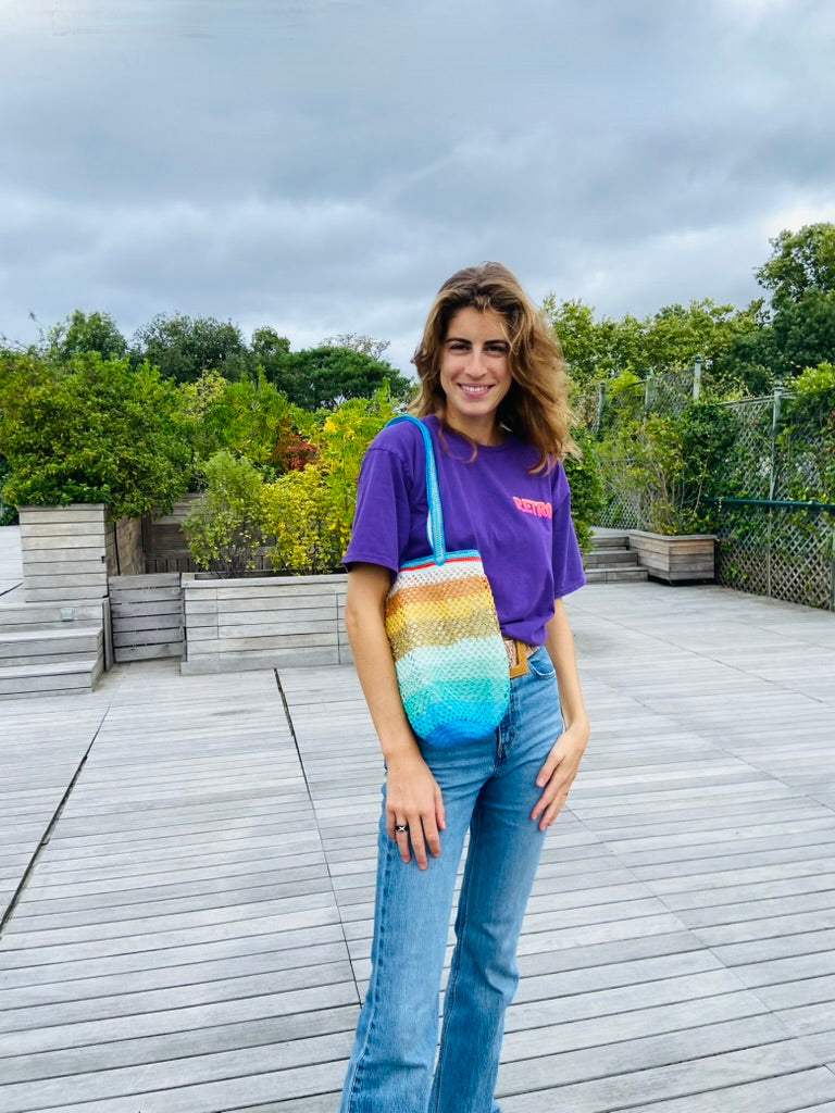 Woman standing on a wooden deck wearing a purple top and jeans, holding an anabaum sirena og bag,  rainbow gradient crochet shoulder bag